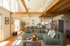 A spacious living area with a green sectional sofa, wooden beams, a dining area, and a kitchen with modern appliances.