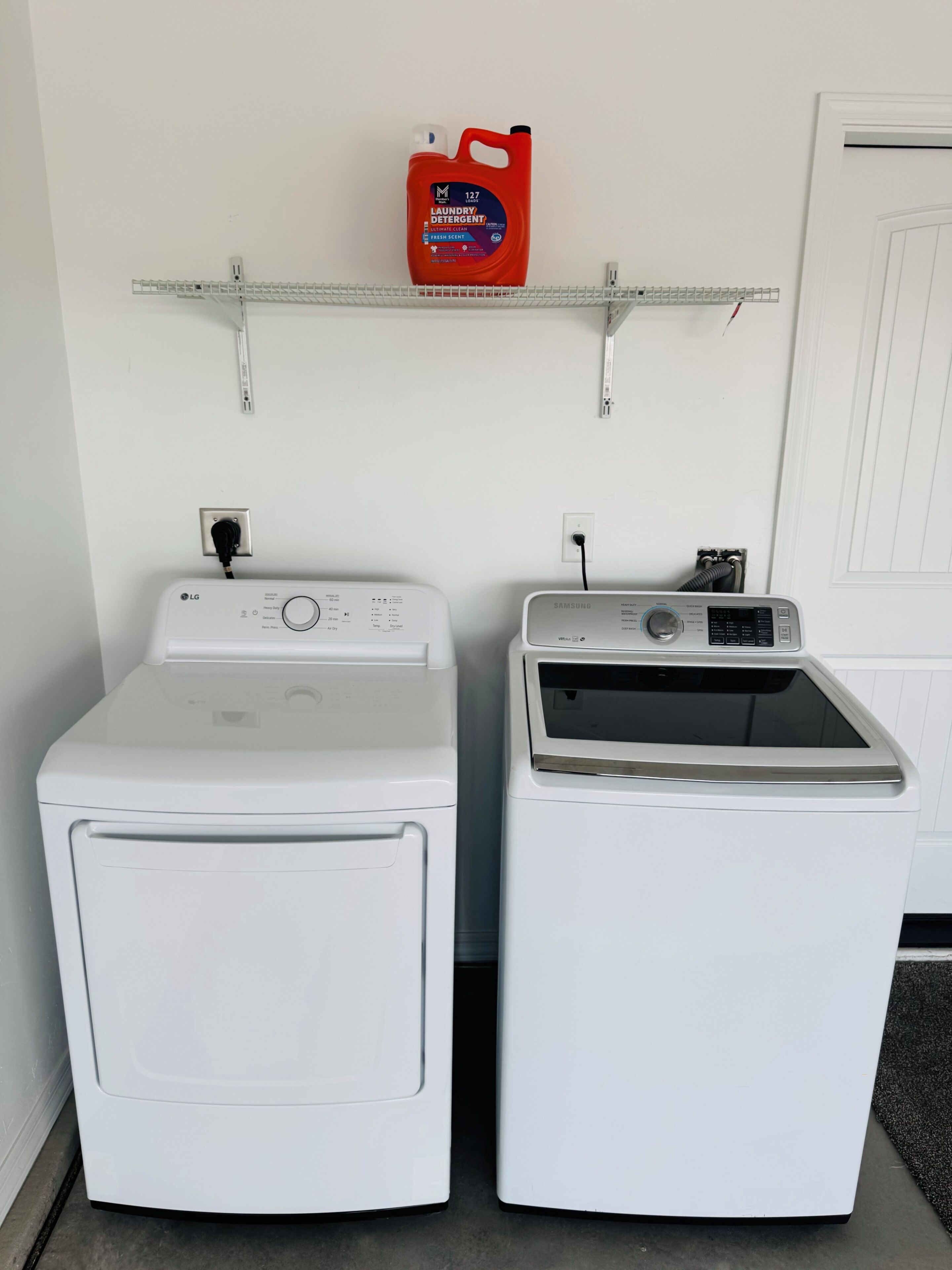 Two white front-loading washing machines side by side, a red detergent bottle on a shelf above, and a door in the background.
