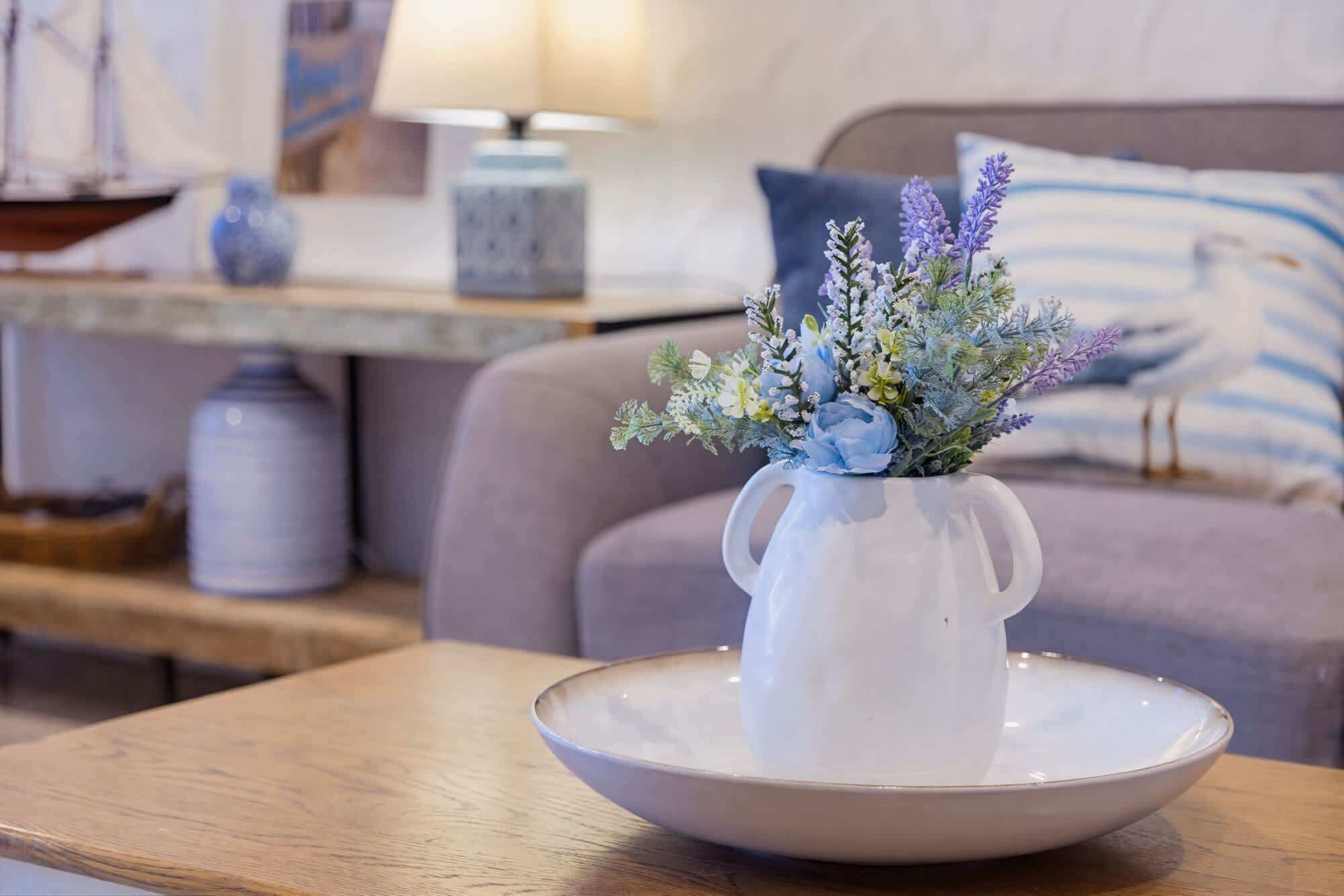 A white ceramic pitcher with a floral arrangement on a white plate, placed on a wooden table in a room with a sofa and a lamp in the background.
