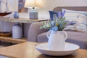 Un pichet en céramique blanche, orné d’un arrangement floral, est posé sur une assiette blanche, elle-même placée sur une table en bois dans une pièce où l’on aperçoit un canapé et une lampe en arrière-plan.