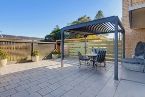Una zona de pati coberta amb taula i cadires, una estructura de pergola i una paret de maó.