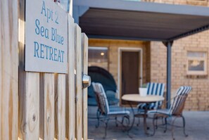 Ein Holzzaun mit einem Schild für einen Rückzugsort, eine überdachte Terrasse mit gestreiften Stühlen und einem Tisch sowie ein Backsteinhaus im Hintergrund.