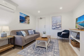 A modern living room with a sofa, coffee table, and armchair, a flat-screen TV, and wall art depicting coastal scenes. - Sea Blue Retreat 2-Bed Unit in Glenelg North (Glenelg North)