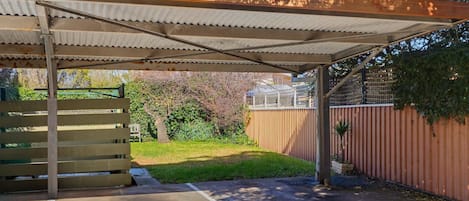 Un espacio al aire libre techado, con piso de concreto, un banco de madera y una cerca de metal.