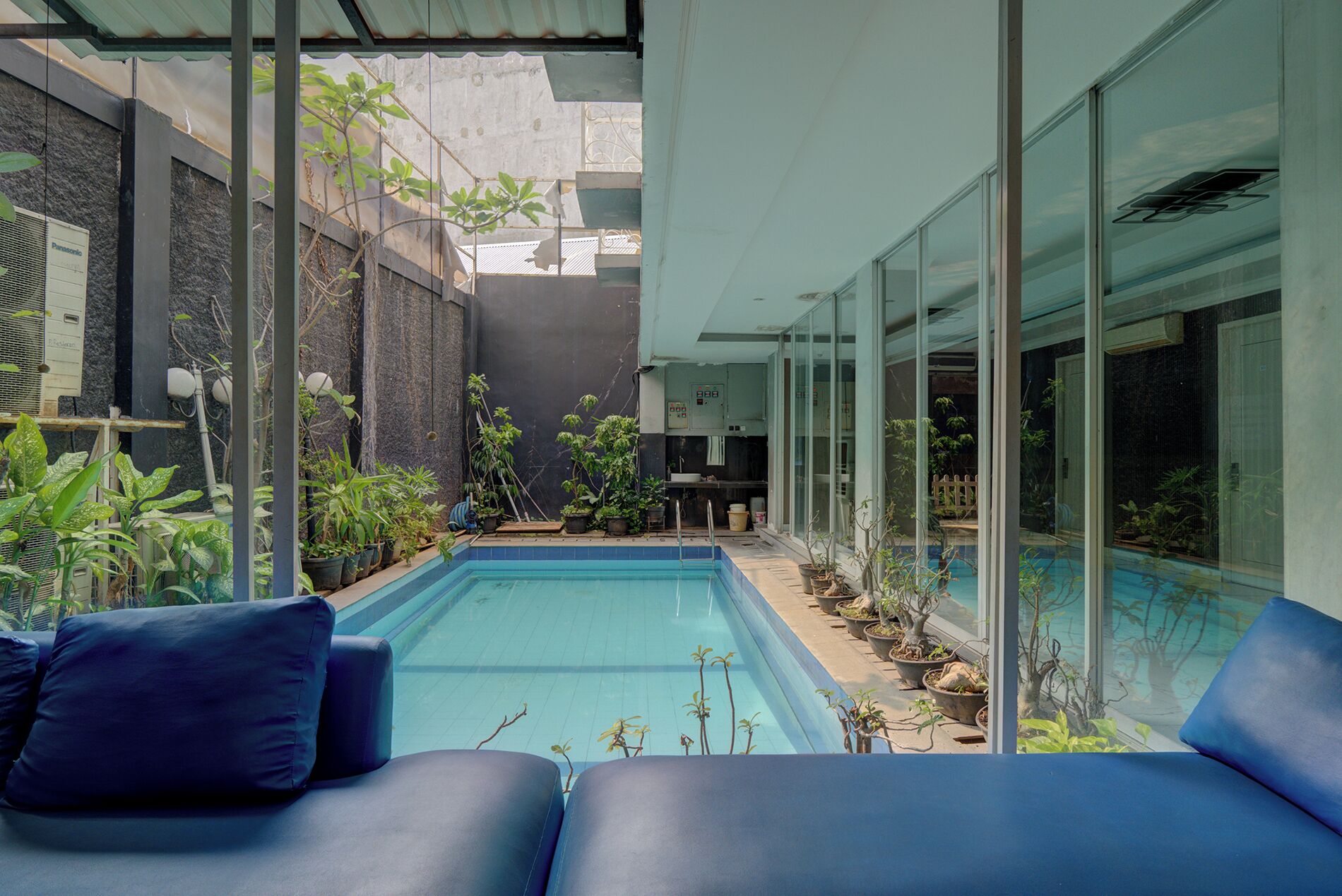 A modern pool area with blue seating, potted plants, and a glass-enclosed kitchen.