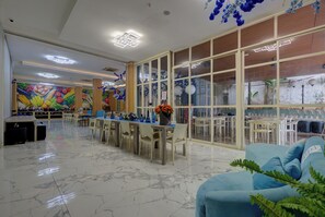 En rymlig hotellobby med ett marmorgolv, en central matsal med bord och stolar samt ett stort fönster prytt med dekorativa blå blommor.