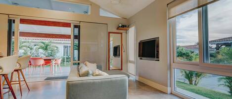A modern living room with a sofa, dining area, and large windows offering a view of the outdoors.