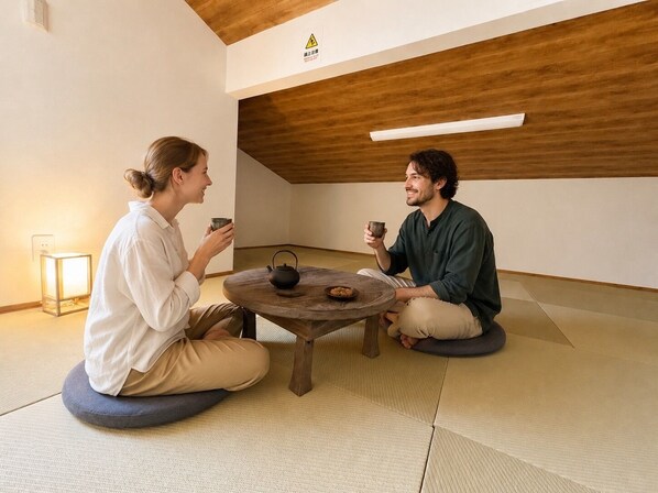 Two people sitting on tatami mats around a low wooden table with a teapot and cups, in a room with a wooden ceiling and a caution sign.