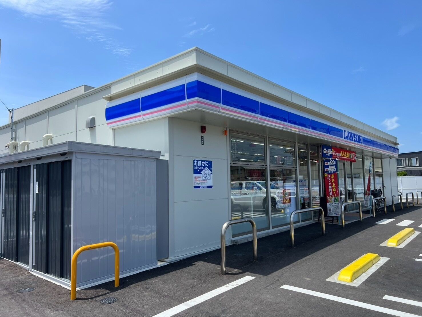 A Lawson convenience store with a clear entrance, a parking lot with marked spaces, and a small enclosed area to the left.
