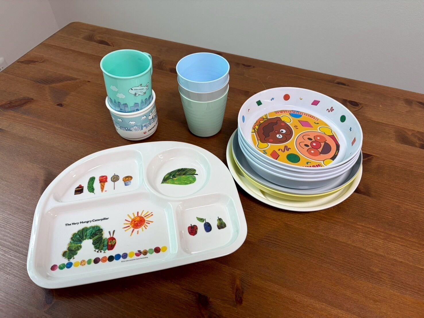 A table with a plate featuring a caterpillar design, a stack of colorful plates with animal prints, and two teal cups.