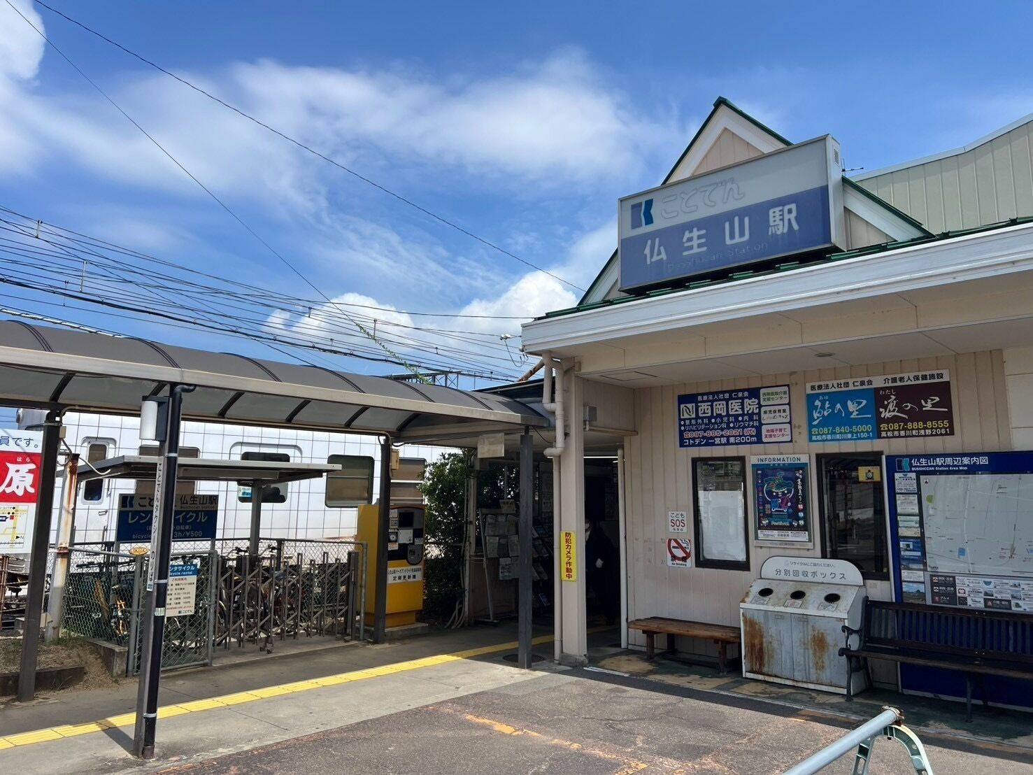 A train station with a covered waiting area, a ticket office, and a small building with multiple signs.