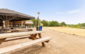 An outdoor seating area with wooden benches and tables, a covered section with a black awning, and a view of a green playground and water body in the background. - 2 bedroom cozy home in Erm (Erm)