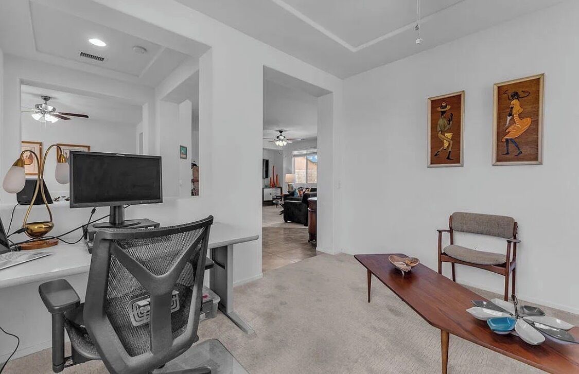 A modern office space with a desk, chair, and computer monitor. There are two framed artworks on the wall and a dining table with plates.