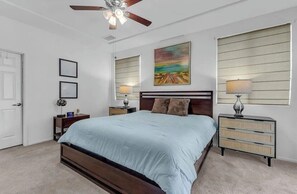 Ein Schlafzimmer mit einem Bett, zwei Nachttischen, einem Deckenventilator, einem Gemälde und einem Fenster mit Jalousien.