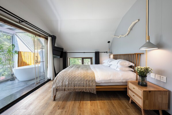 A modern bedroom with a large bed, a wooden nightstand, a bedside table, a hanging light fixture, and a view of the outside.