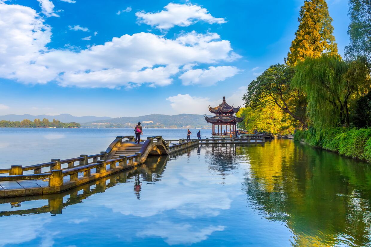 A serene lakeside scene with a traditional pavilion, a wooden bridge, and a clear sky.