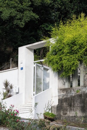 Un edificio blanco con una reja, rodeado de vegetación y un muro de concreto.