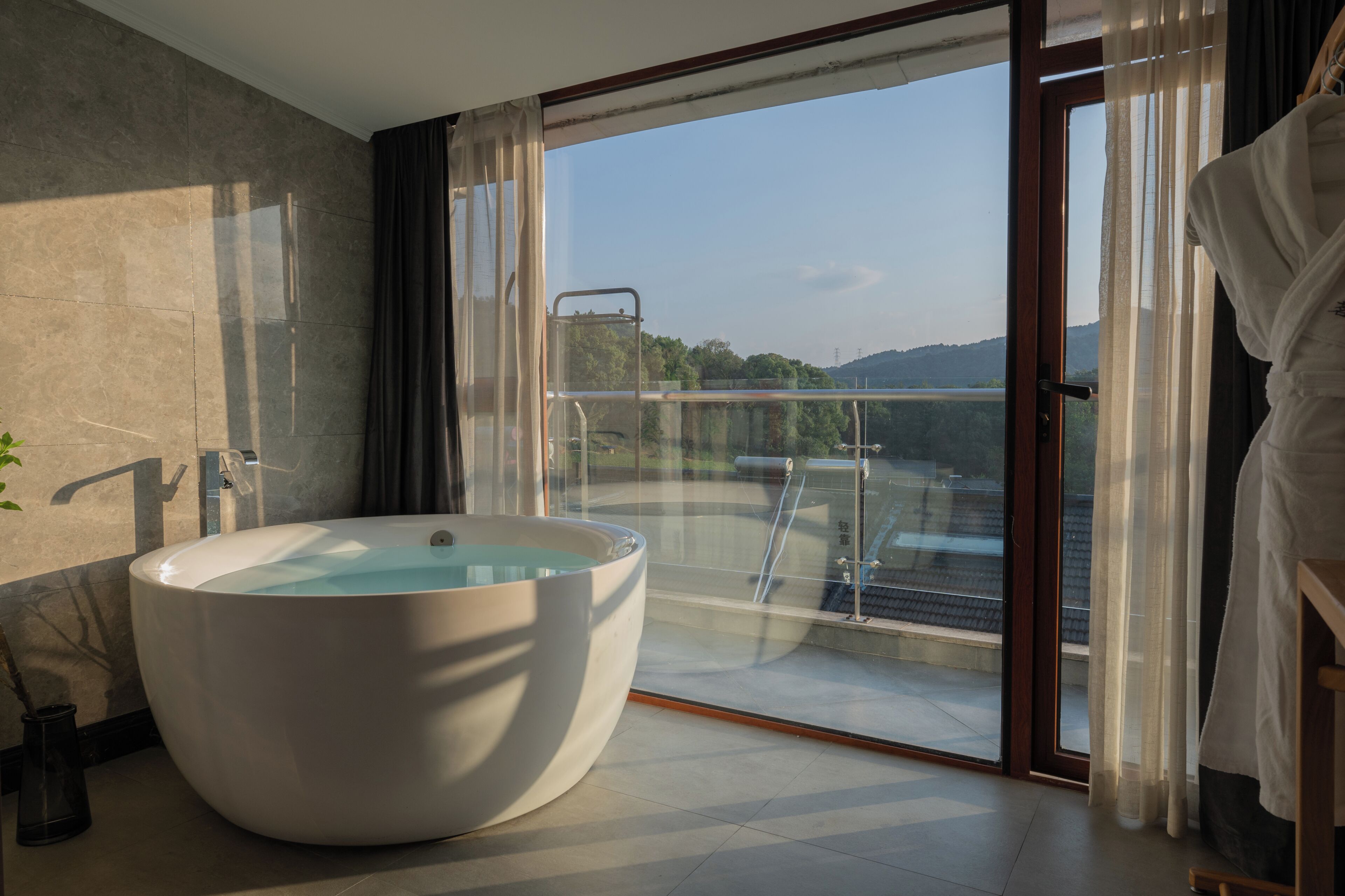 A modern bathroom with a large freestanding bathtub, a glass-enclosed balcony with a view of the landscape, and a wooden door.