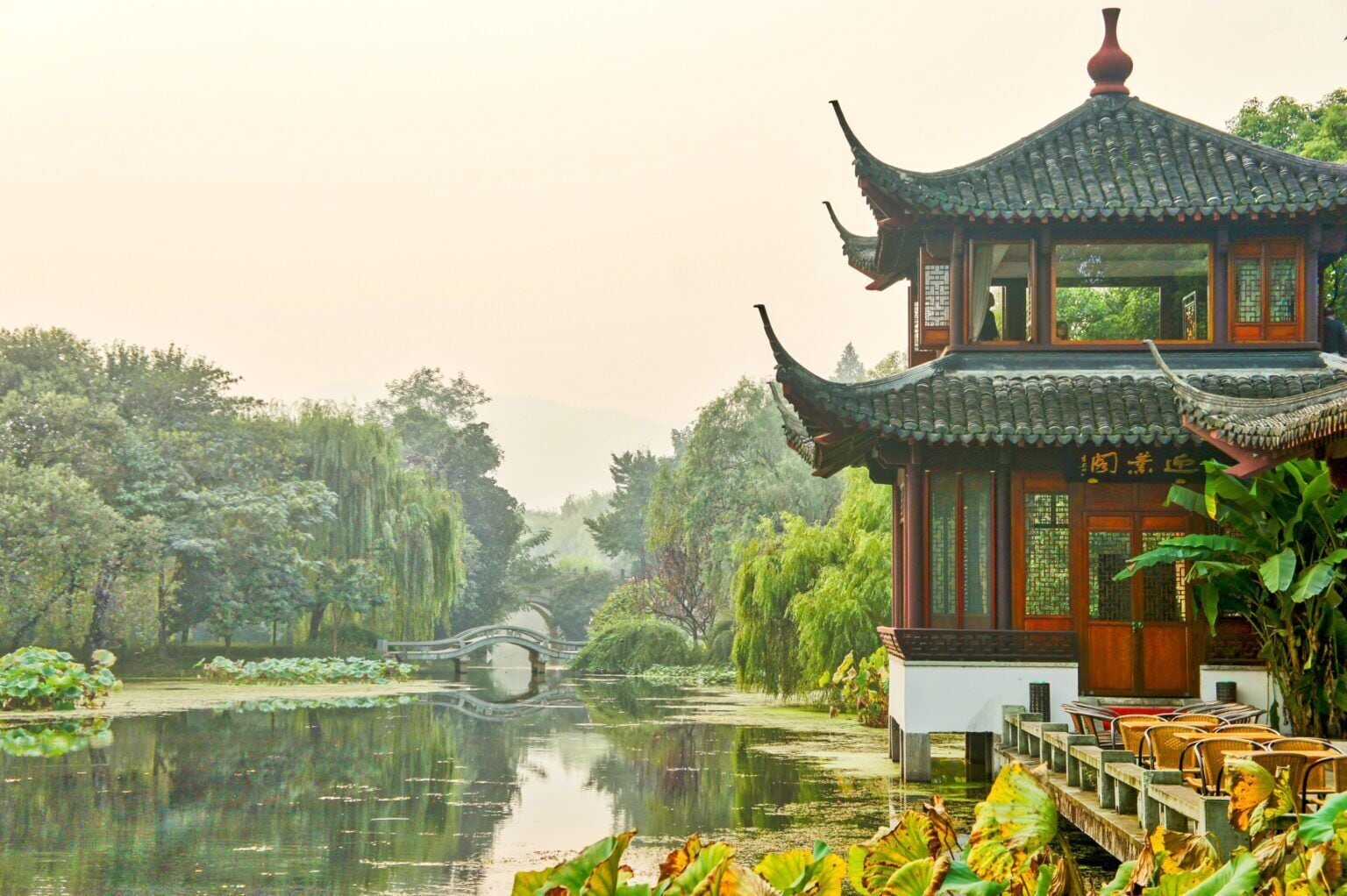 A traditional Chinese pavilion by a tranquil pond with a bridge in the background.