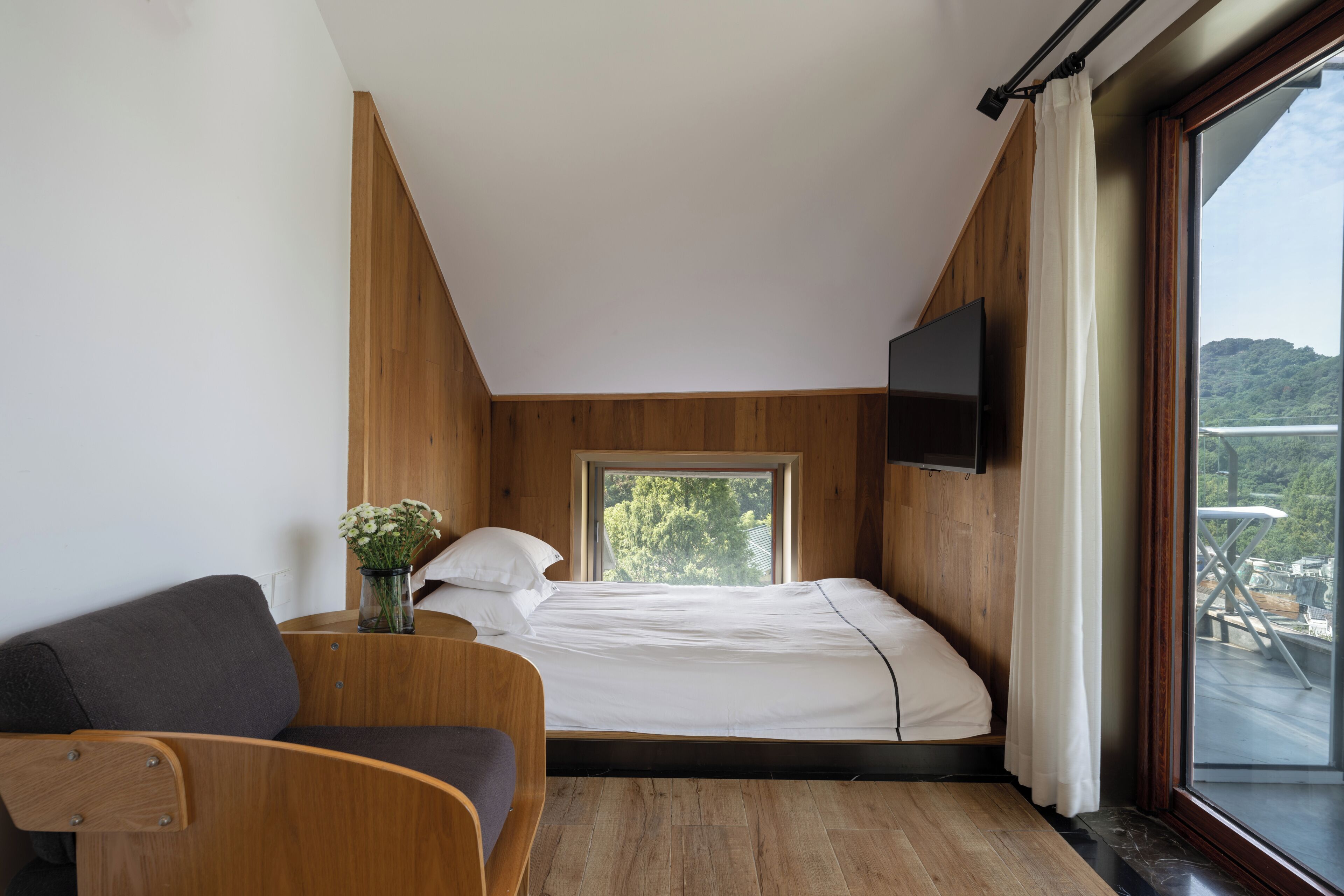 Een slaapkamer met een bed, een aan de muur bevestigde televisie, een raam met uitzicht op groen en een houten vloer.