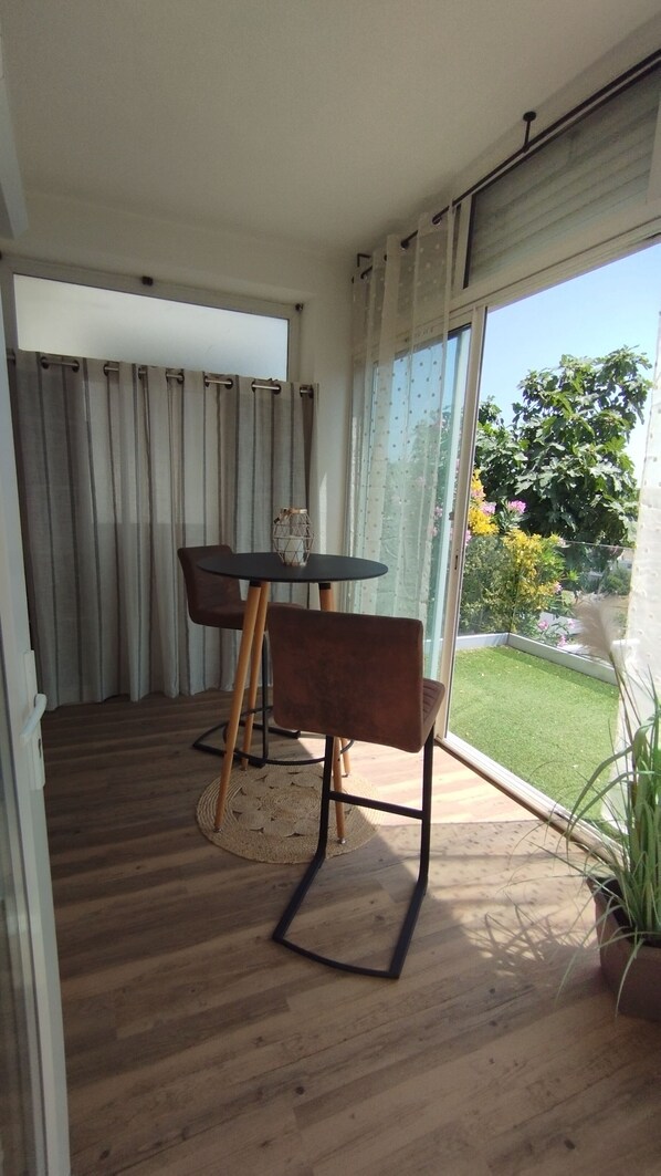 Een kamer met een houten vloer, een ronde tafel met twee stoelen, een plant in pot en uitzicht op een tuin door een glazen deur.