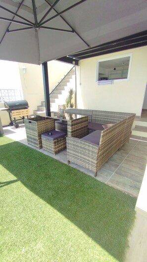 Een terras buiten met rieten meubels, een barbecue en een overdekt gedeelte.