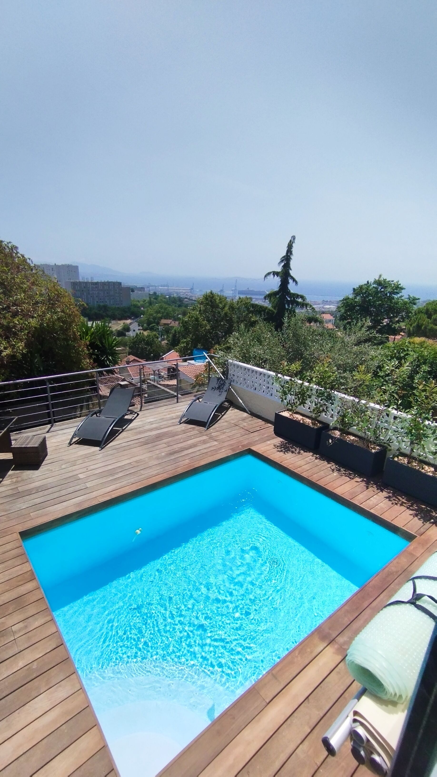 Une piscine rectangulaire avec une terrasse en bois, entourée d’une clôture blanche, et offrant une vue sur le paysage urbain lointain.