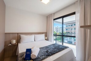A hotel room with a large bed, a nightstand, a lamp, a window with a view of buildings, and a blue booklet on the bed. - Modern Apartment in a Cond. With gym Hgr0101 (Salvador)