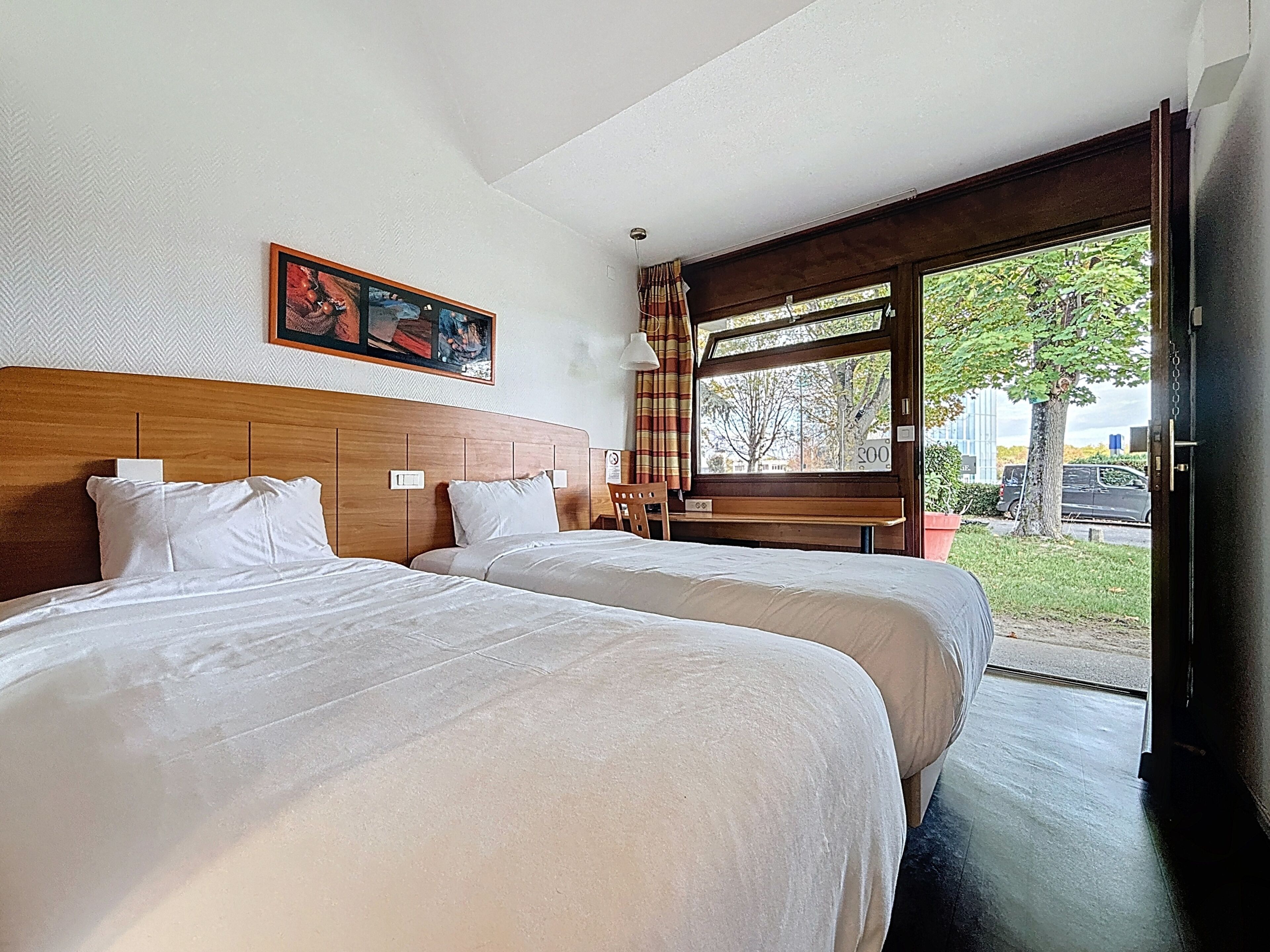 A hotel room with two beds, a wooden headboard, a bench, and a large window with a view of the outdoors.