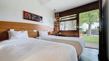 A hotel room with two beds, a wooden headboard, a bench, and a large window with a view of the outdoors.