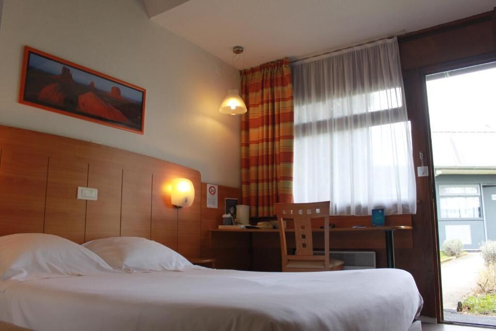 A hotel room with a bed, a desk, a chair, a painting on the wall, and a window with curtains.