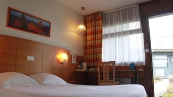 A hotel room with a bed, a desk, a chair, a painting on the wall, and a window with curtains.