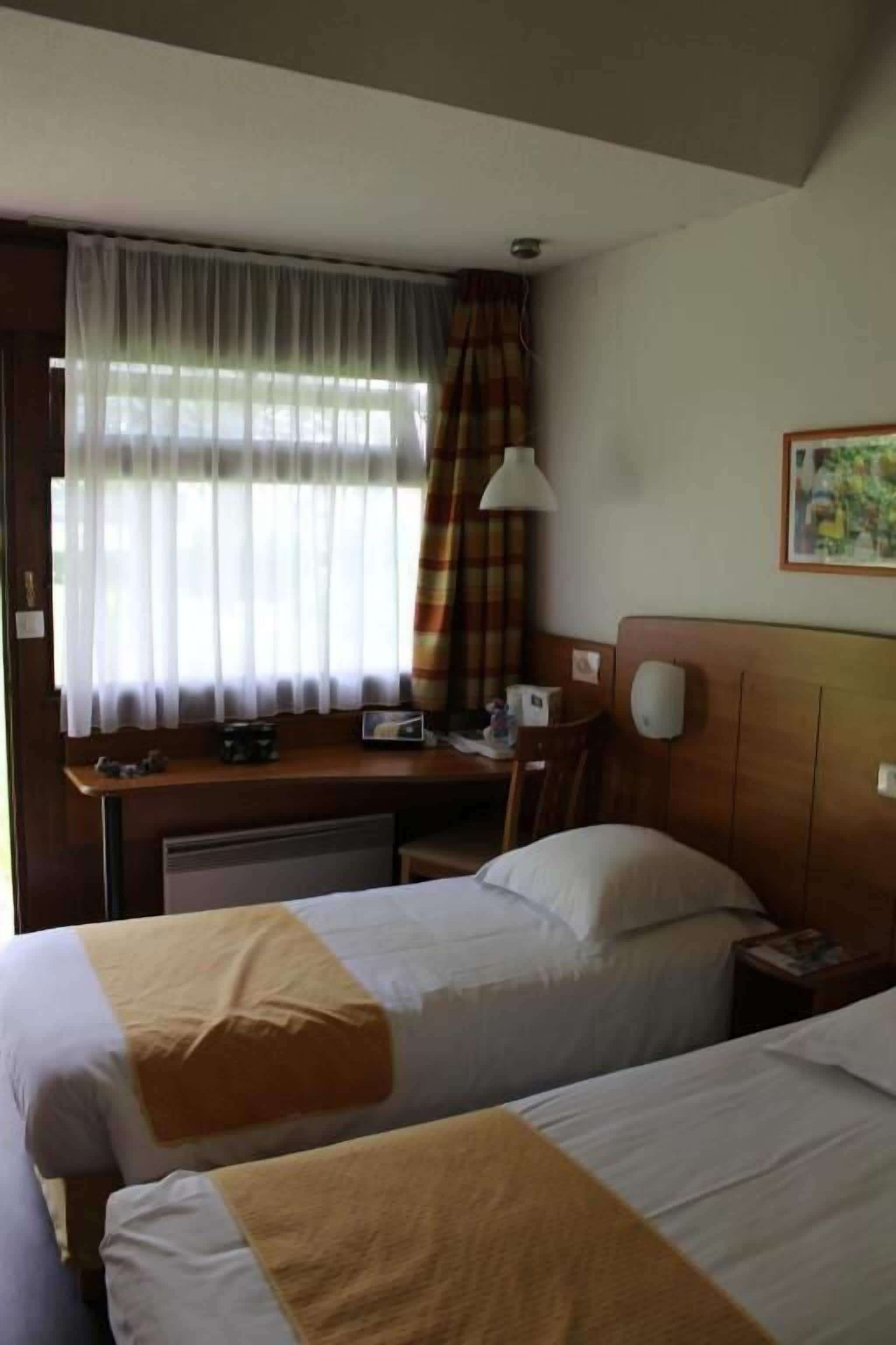 A hotel room with two beds, a desk, a chair, a lamp, and a window with curtains.