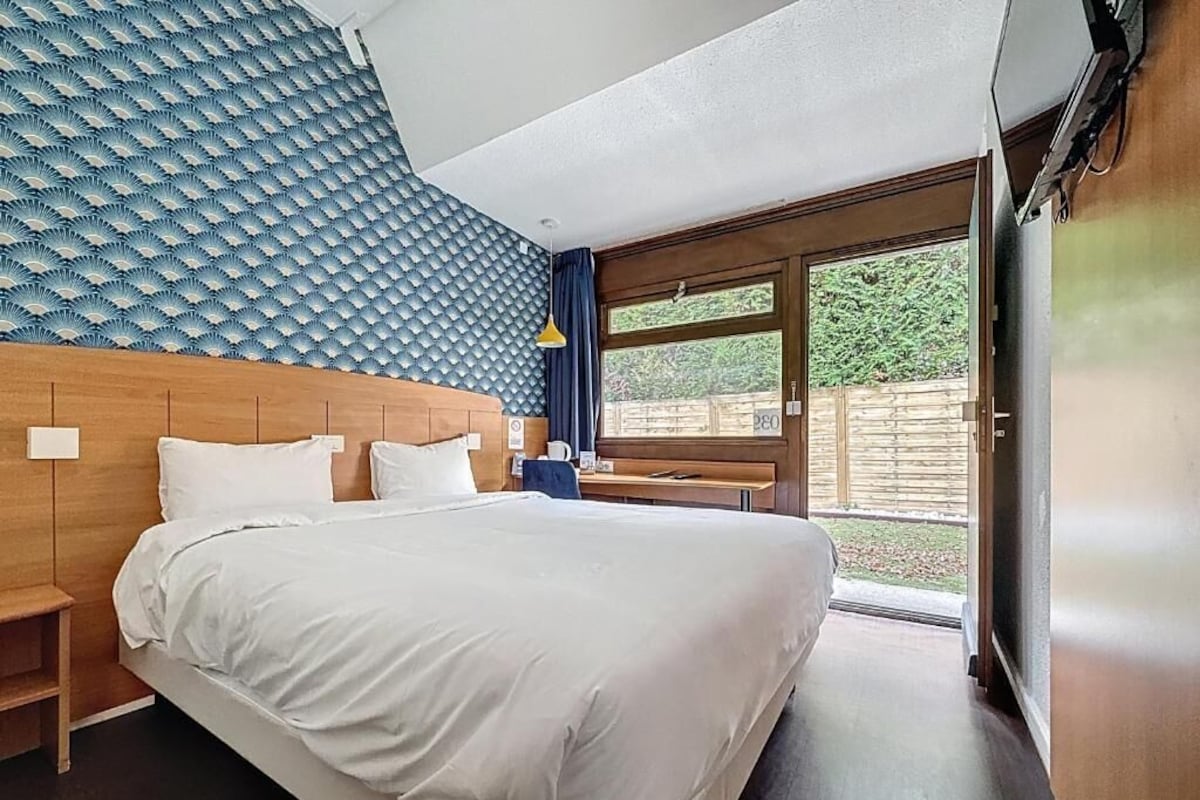 A modern hotel room with a large bed, a wooden headboard, a window with a view of greenery, and a wall with a textured pattern.