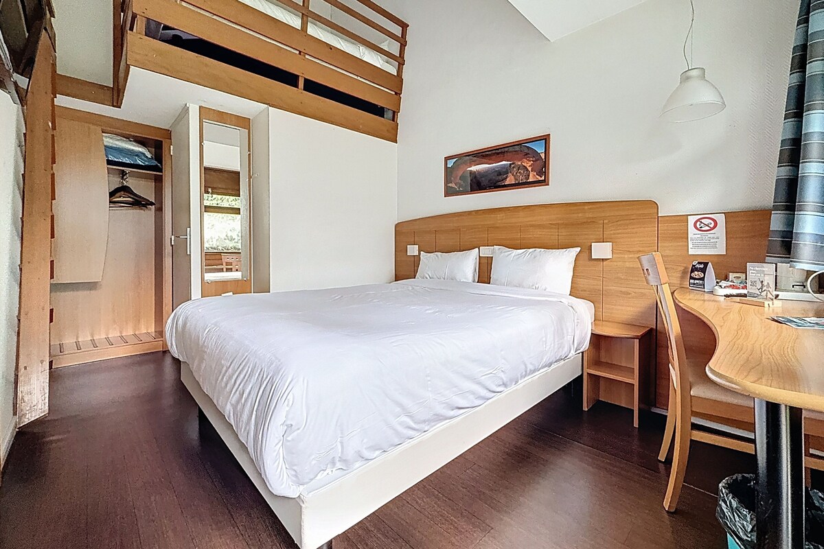 A hotel room with a bunk bed, a desk with a chair, a hanging light fixture, and a framed picture on the wall.
