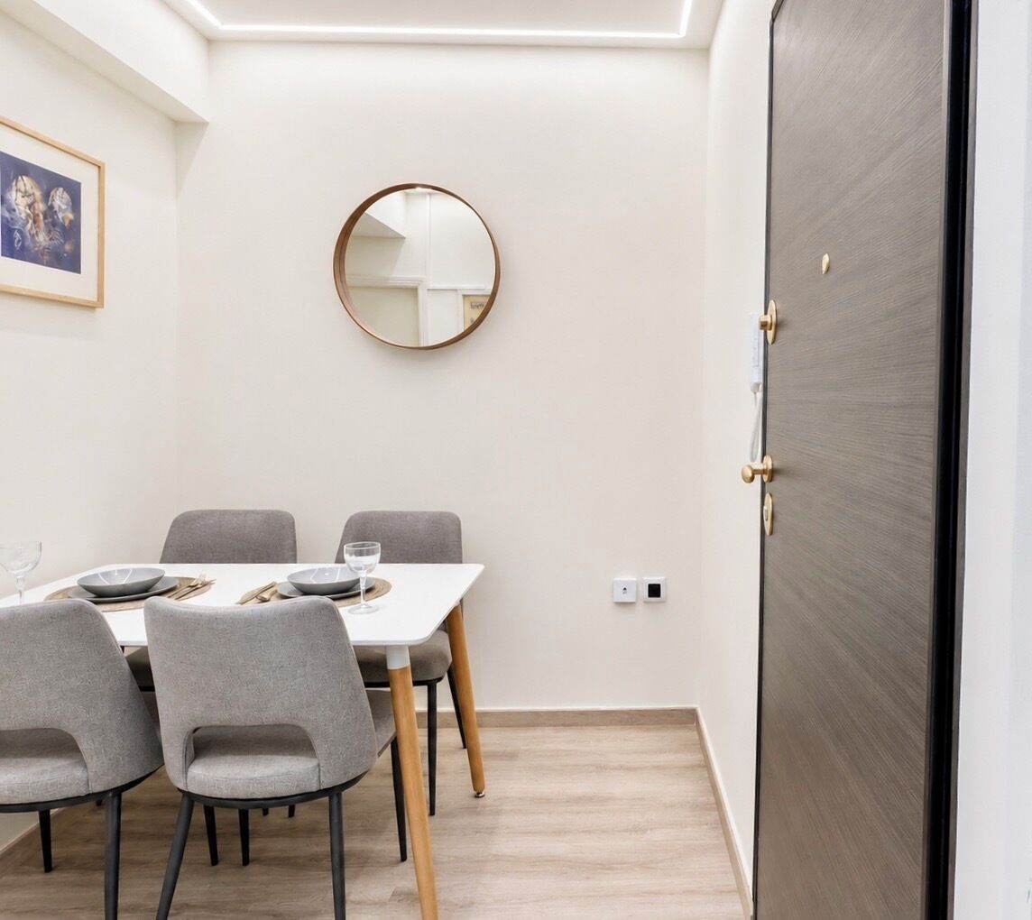 A dining area with a table set for four, grey chairs, a round mirror on the wall, and a dark wooden door.