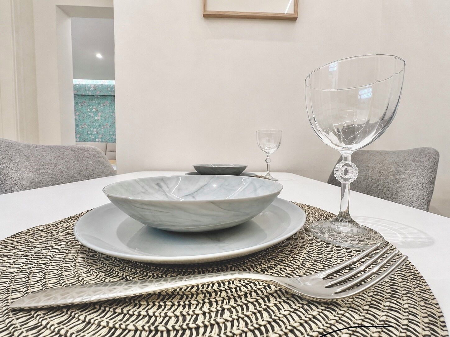 Una mesa para comer con un bol, plato, copa de vino y cubiertos sobre un posavasos tejido.