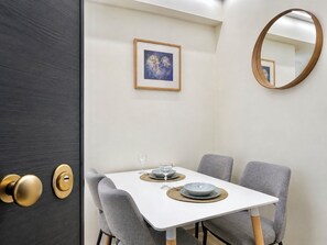 A dining area with a white table, grey upholstered chairs, a framed artwork on the wall, and a round mirror. - Riviera Escape - Stylish 1BR Near the Sea by ΜΡS (Voula)