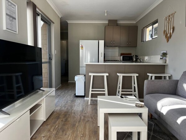 Un espacio habitable compacto con kitchenette, una zona de estar con sofá y taburetes, y una televisión.