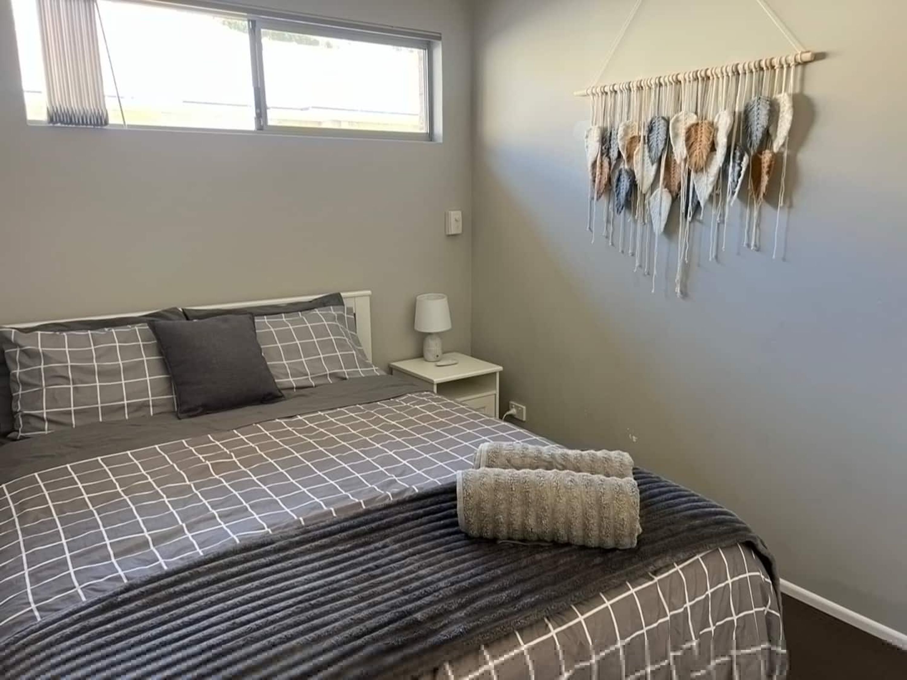 Un dormitorio con cama, almohadas, una mesita de noche, una lámpara y un colgador de macramé en la pared.
