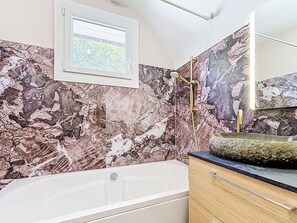Ein Badezimmer mit Badewanne, einer Dusche mit goldener Duschbrause, einem Waschbecken mit Marmorplatte und einem Fenster mit Blick ins Grüne.
