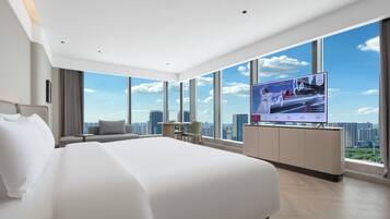 A modern hotel room with a large bed, a TV mounted on the wall, and a city view through large windows.