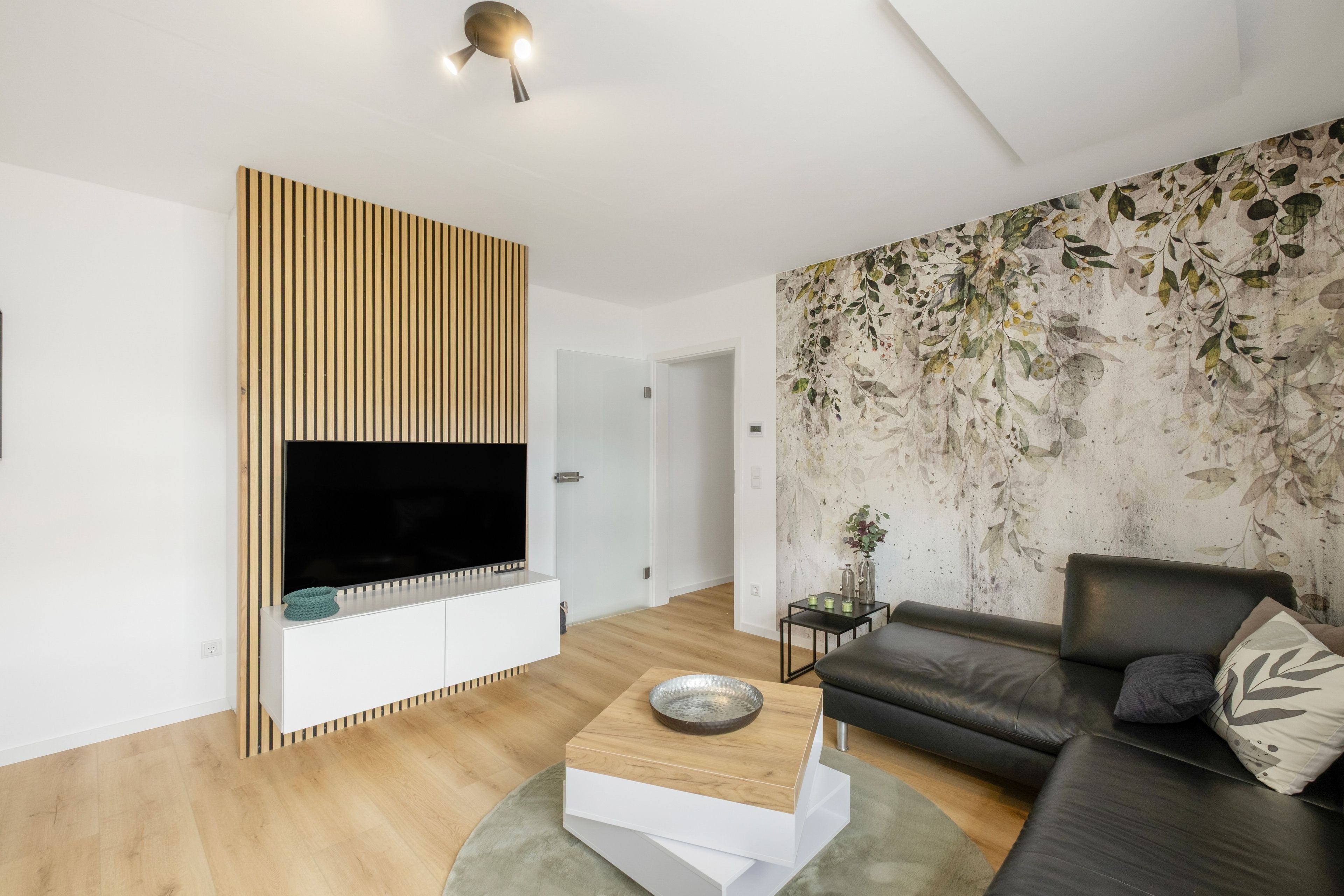Ein modernes Wohnzimmer mit einem flachbildfernseher an einer Holzvertäfelung, einem schwarzen Ledersofa und einer Wand mit einem Blattmuster.