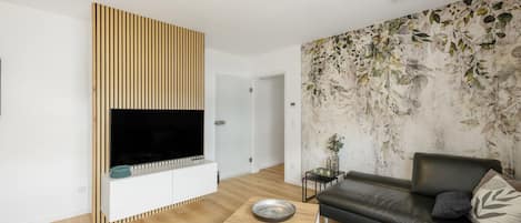 A modern living room with a flat-screen TV mounted on a wooden panel, a black leather sofa, and a decorative wall with a leaf pattern.