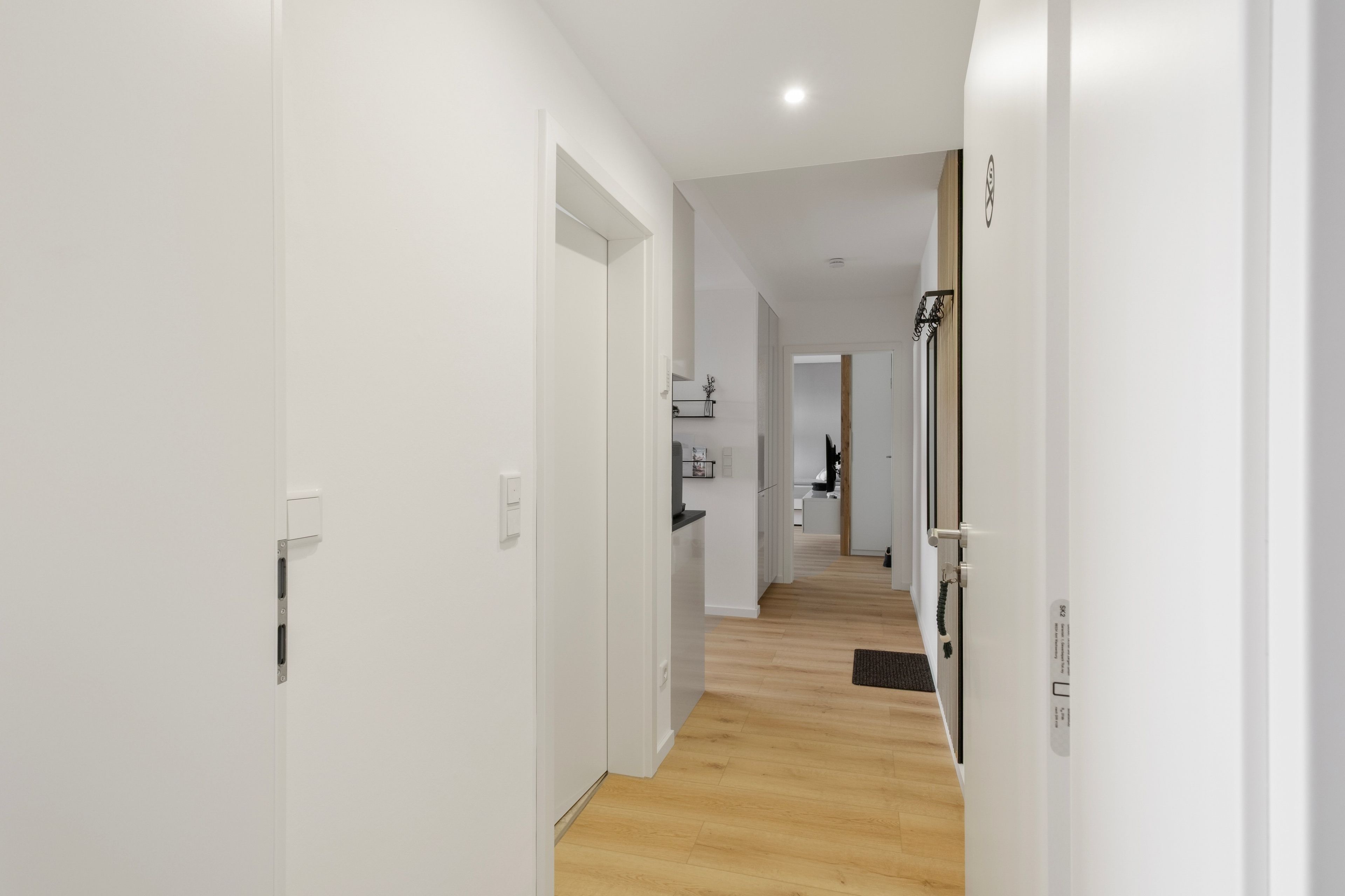A corridor with white walls and wooden flooring leading to a kitchen area.