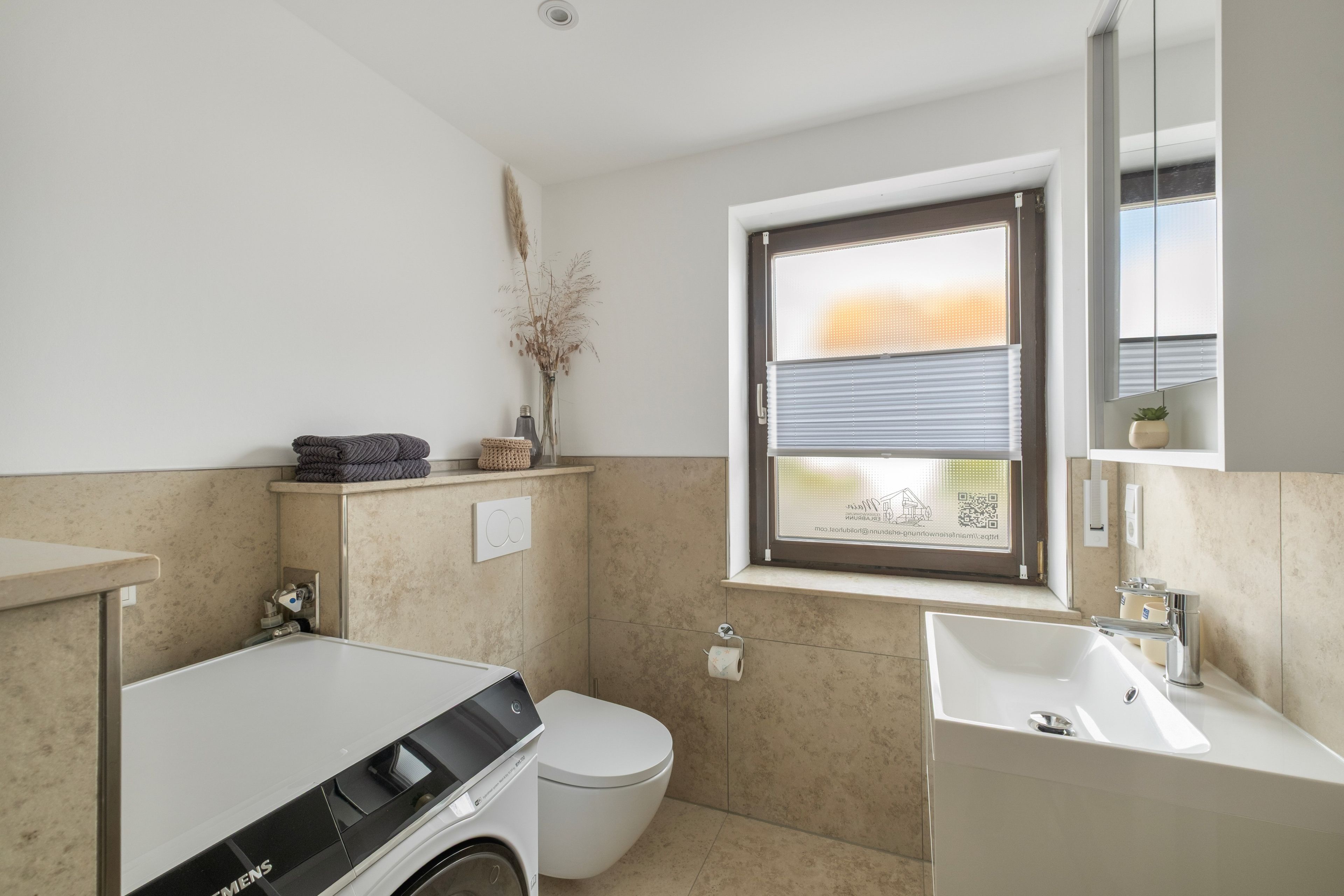 Een moderne badkamer met een wasmachine, toilet, wastafel en een raam met zonwering.