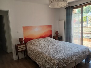A bedroom with a bed, a nightstand, a lamp, a painting, and a view through a window. - Spacious apartment with a south-facing garden and terraces (Le Monêtier-les-Bains)
