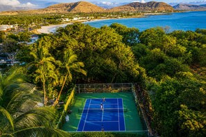 A tennis court surrounded by lush greenery and a beach in the background. - Hawaiian Princess Beachfront 1BR (Waianae)