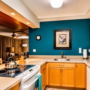 A modern kitchen with a stove, oven, and sink, wooden cabinets, a clock, and a framed artwork on the wall.