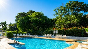 Ein rechteckiger Pool mit klarem, blauem Wasser, umgeben von einer gefliesten Liegefläche. Weiße Liegestühle säumen den Poolrand, und eine Metallleiter führt ins Wasser. Das Areal ist durch einen niedrigen Zaun begrenzt, im Hintergrund sind Bäume und Grün anzusehen.